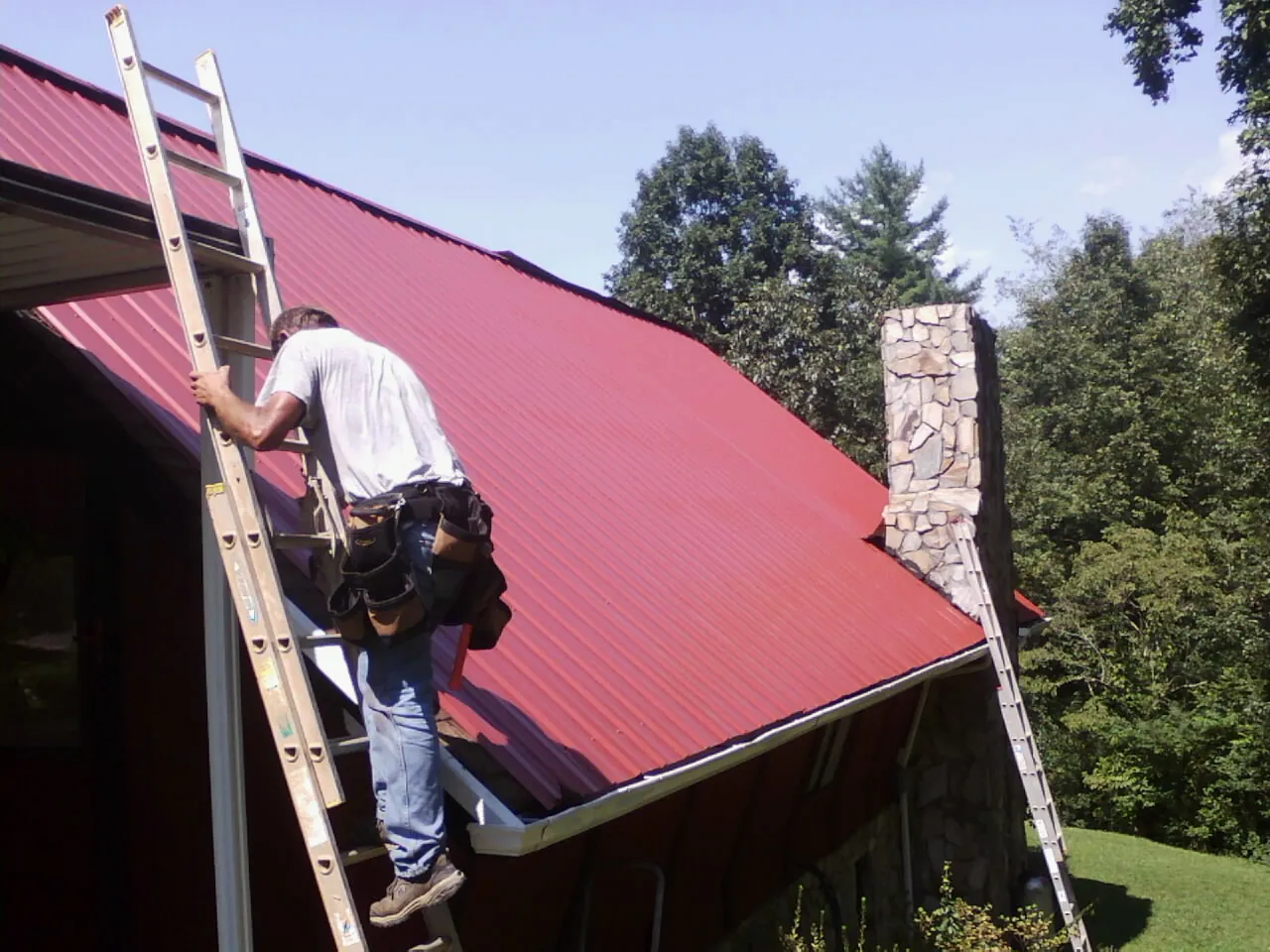 Roofer on a ladder beside a red metal roof and stone chimney — Soffit & Fascia Repair by Roofing Brothers in Warminster